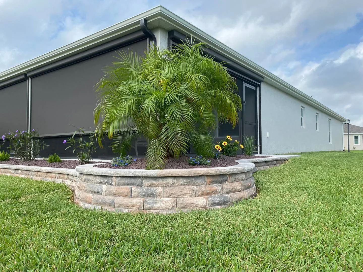 Exterior view: grey house with a stone retaining wall and a palm tree surrounded by greenery.