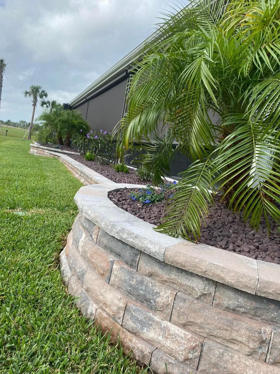 Stone retaining wall bordering a landscaped garden with green grass, palm trees, and a dark-colored building.