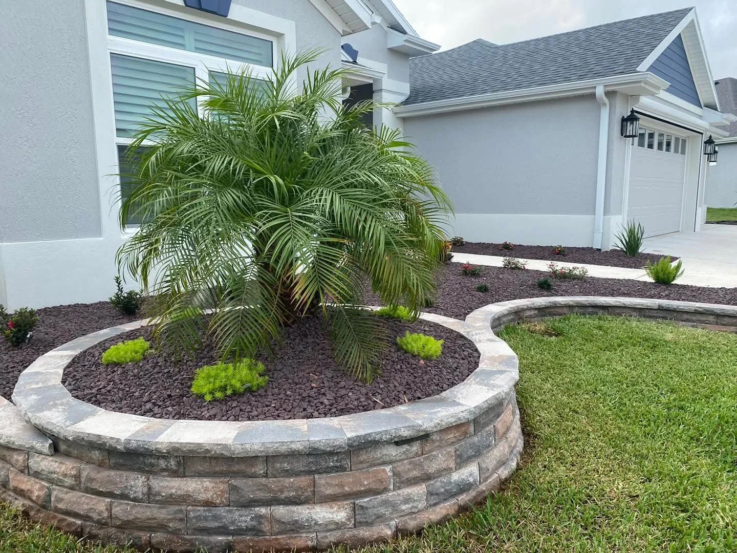 Landscaped front yard with a raised garden bed, palm tree, and brick retaining wall.