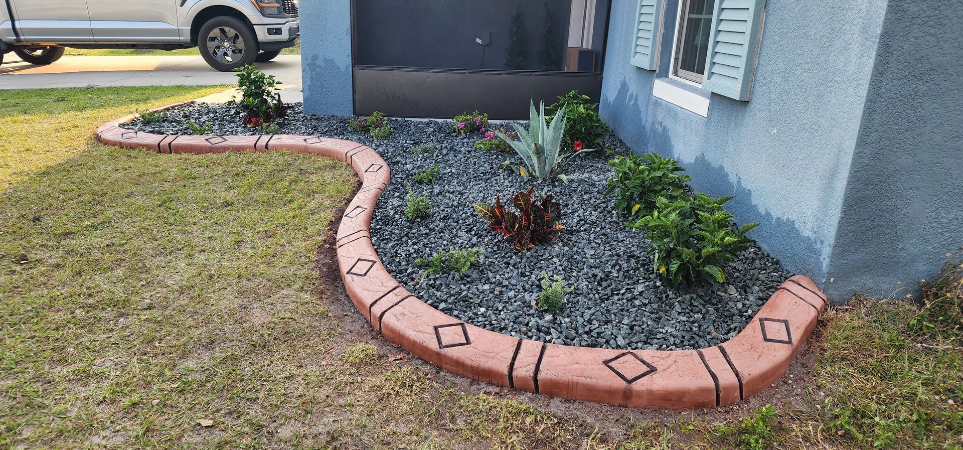 Brick-edged flower bed with dark stones and green plants in front of a blue house.