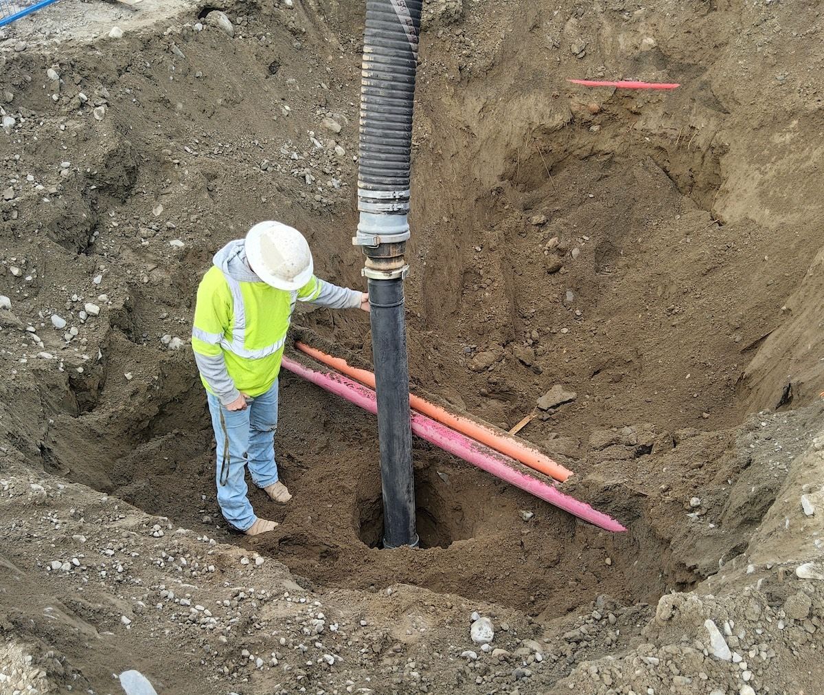 Construction worker in a hole, inspecting a black pipe and coloured conduits. Dirt, hard hat, safety vest — Demetrius Projects in Queanbeyan East, NSW