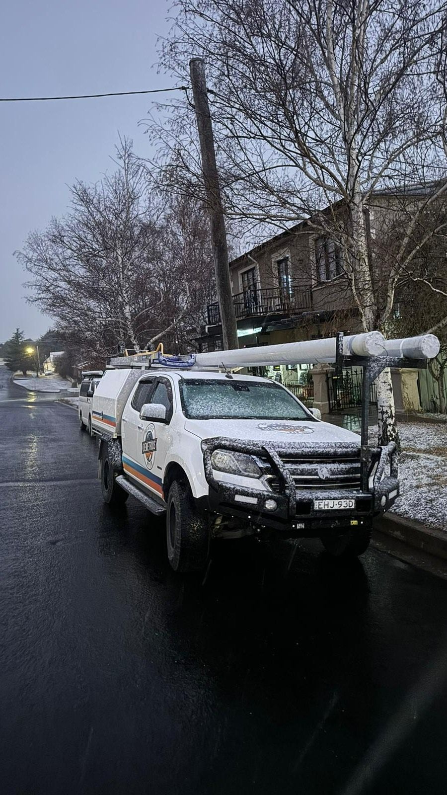 White utility truck with equipment on roof parked on a wet street next to a snow-covered power pole. Building in background. — Demetrius Projects in Queanbeyan East, NSW