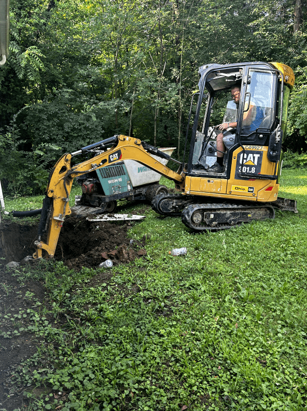 A man is driving a small yellow excavator in a yard.