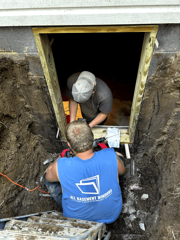 Two men are working on a window in a basement.