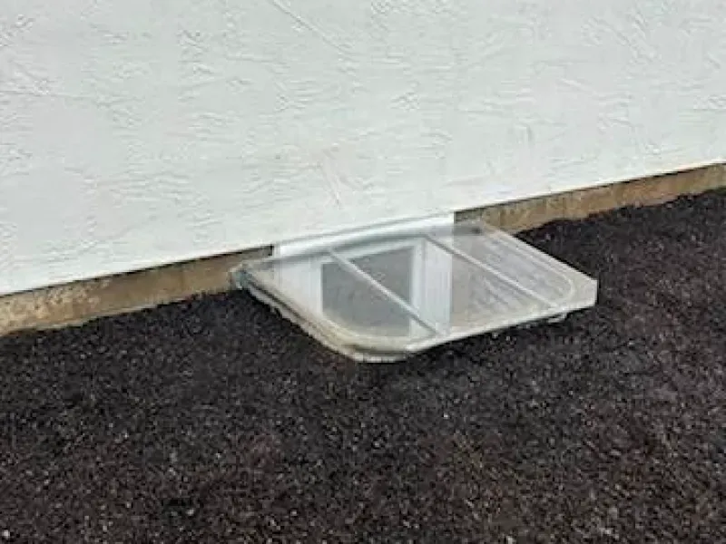 A window well with a clear cover is sitting on the ground next to a wall.