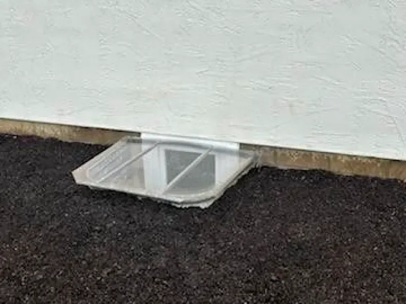 A window well with a cover on it is sitting on the ground next to a wall.