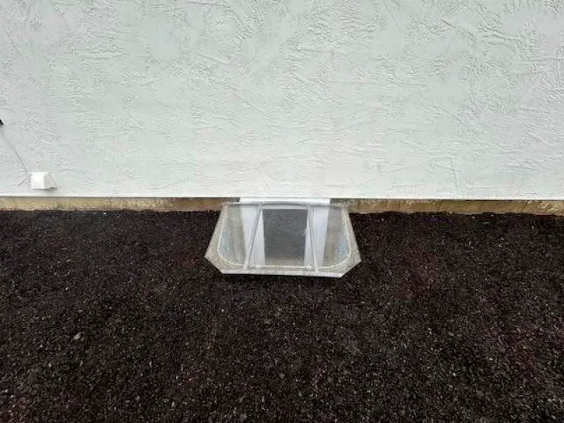 A window well is sitting on top of a pile of dirt next to a wall.
