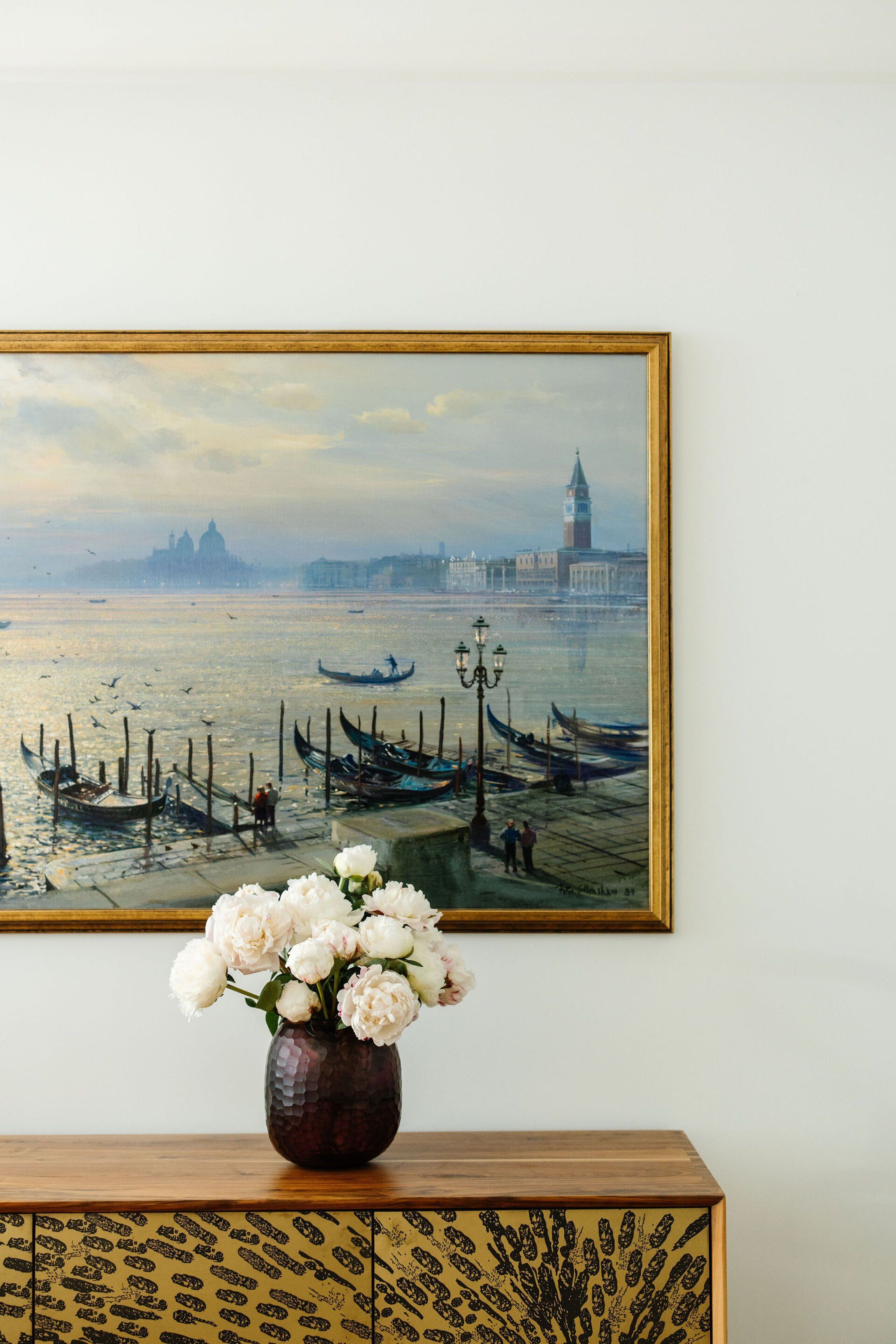 A vase of flowers is sitting on a dresser under a painting of boats in the water.