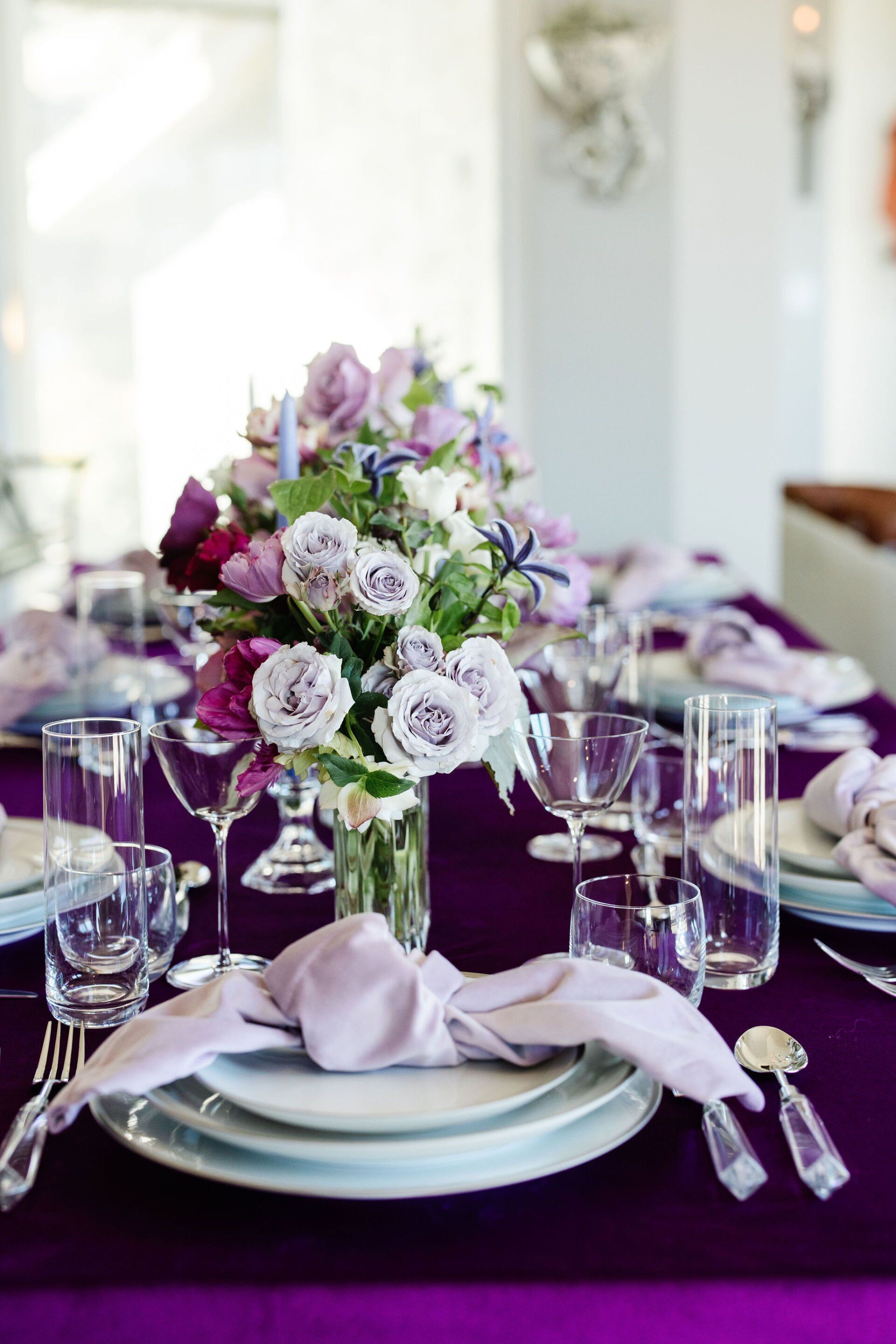 A purple table setting with plates , glasses , napkins and a vase of flowers.