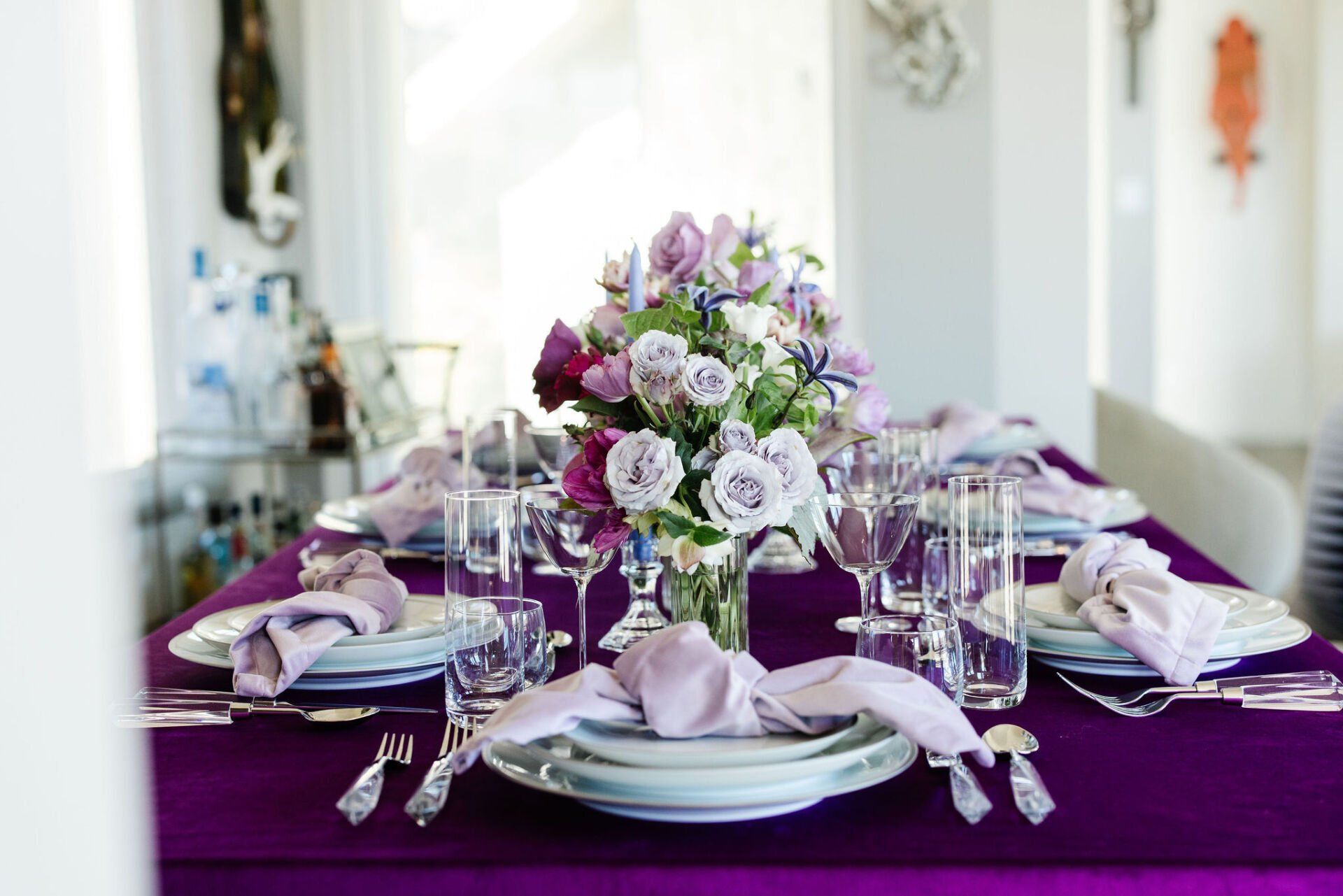 A long purple table with plates , glasses , and a vase of flowers on it.