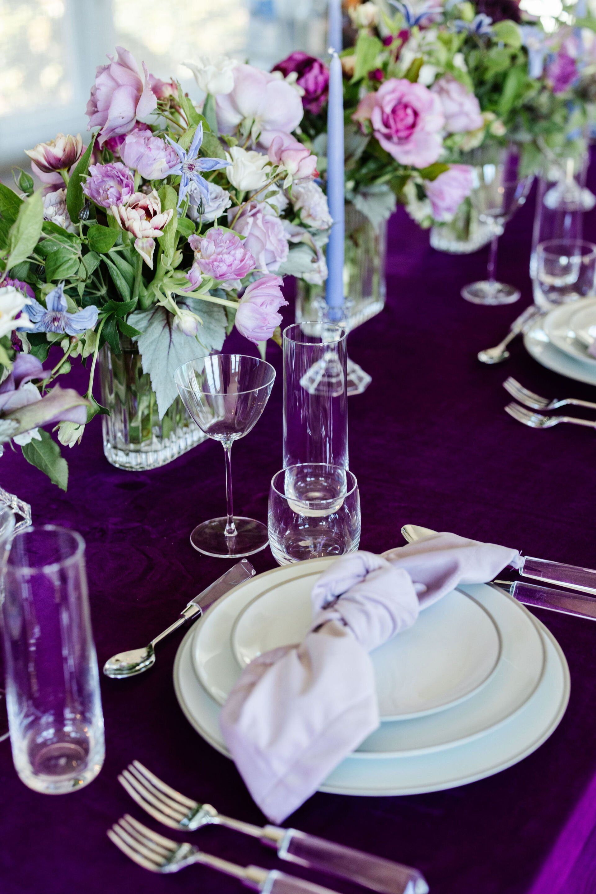 A table setting with plates , silverware , candles and flowers on a purple table cloth.