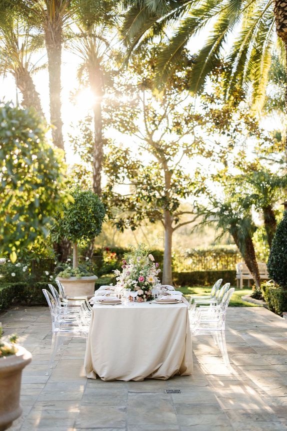 A long table with a white tablecloth is sitting on a patio surrounded by palm trees.