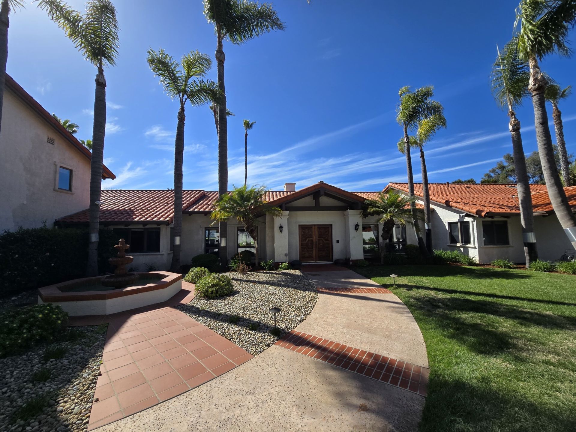 Courtyard of a Spanish-style house with red tile roofs, palm trees, and a curved walkway under a blue sky