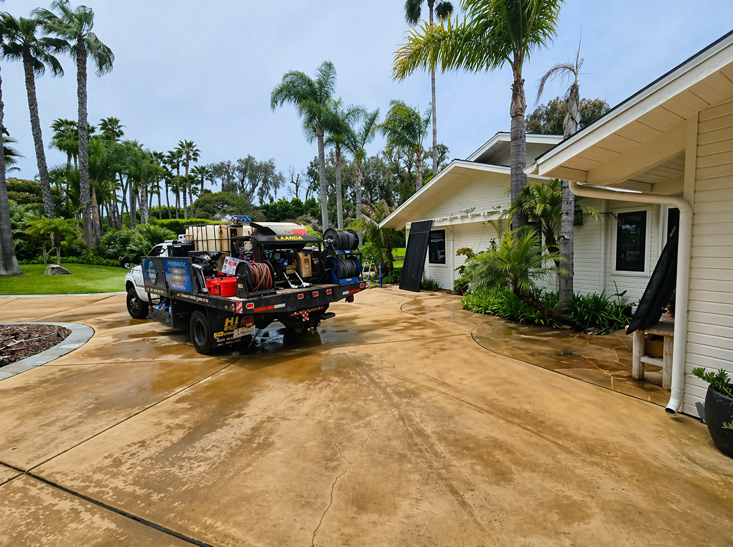 Utility truck on a wet driveway beside a white house, with palm trees and damaged landscaping in the background