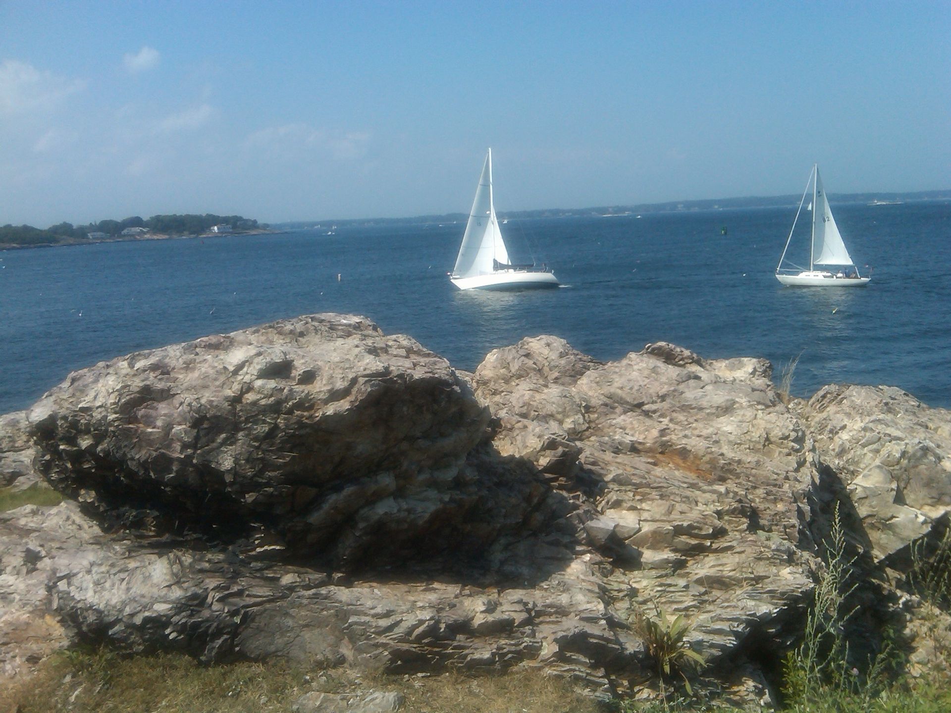 Two sailboats are floating on the water near a rocky shoreline