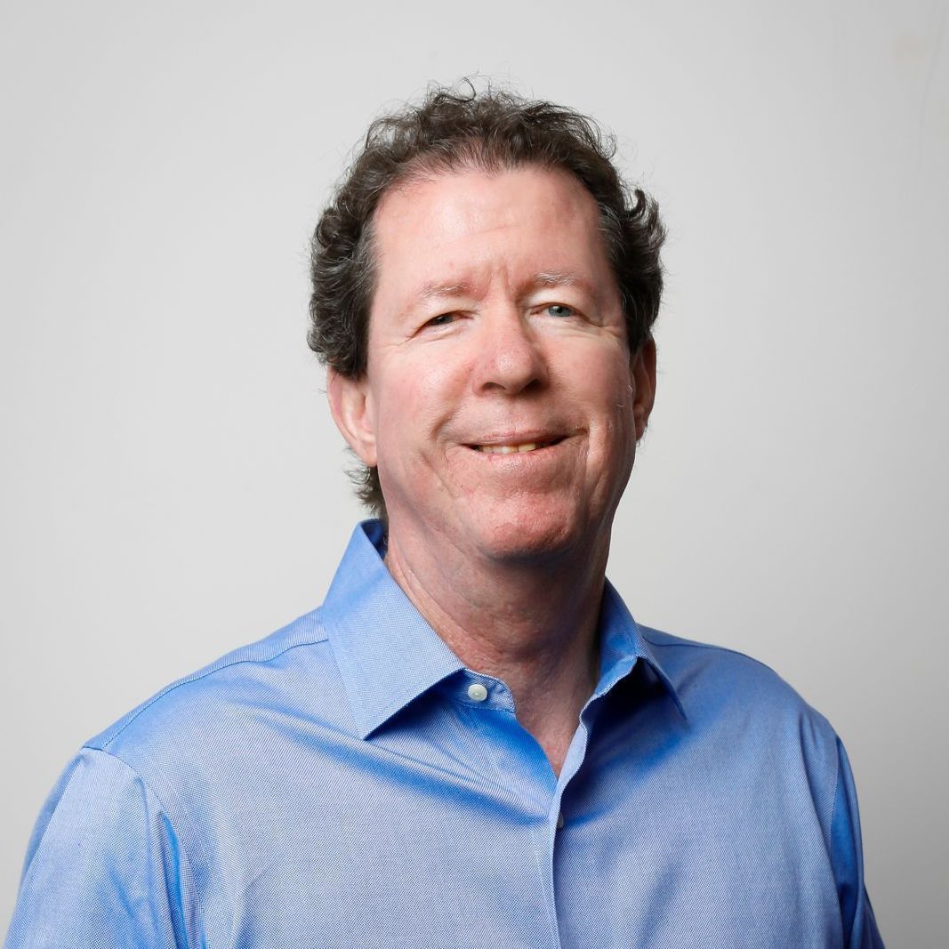 A person with curly hair smiling while wearing a light blue button-down shirt against a plain, light gray background.