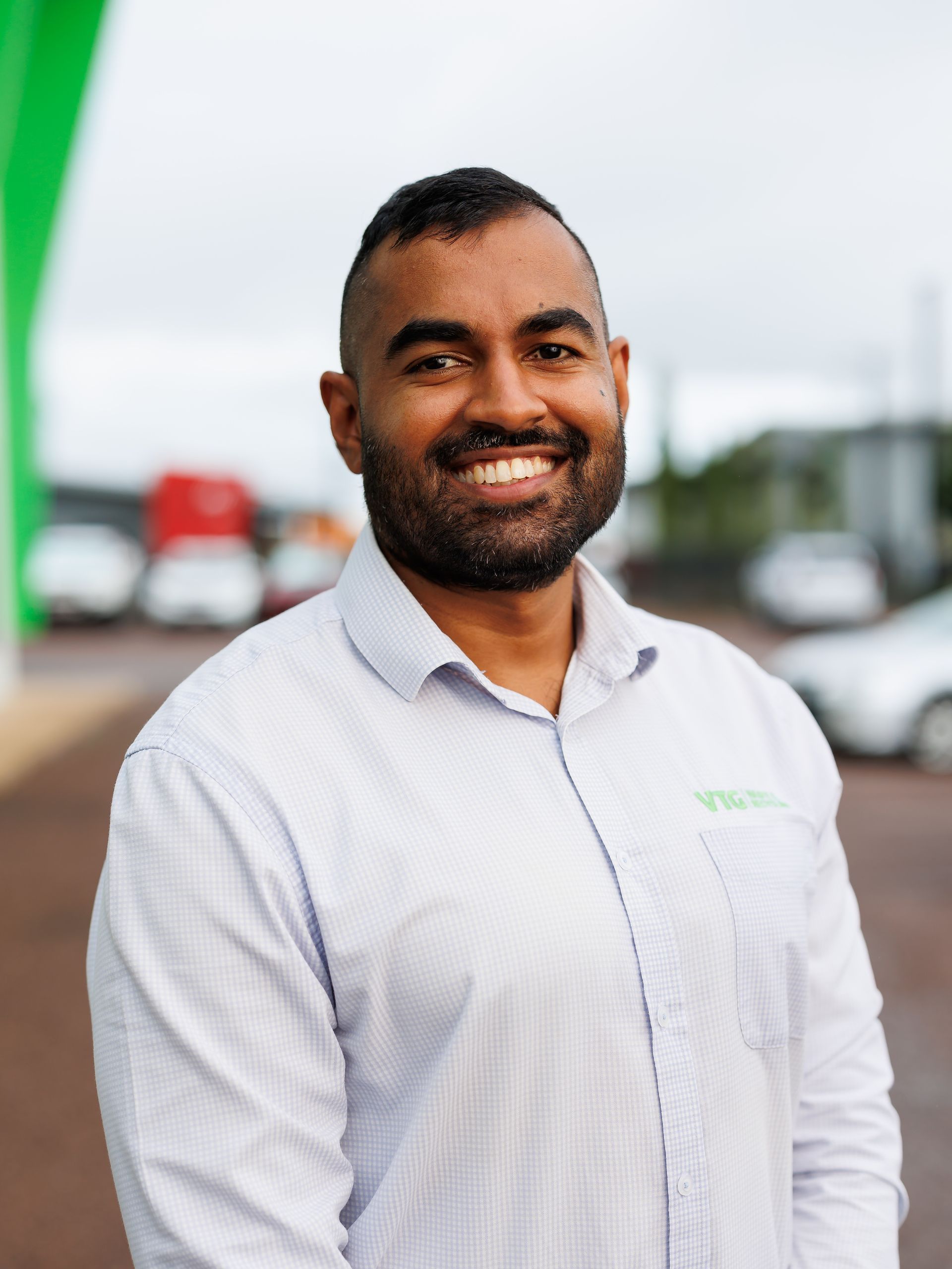 A smiling person in a light blue patterned shirt stands outdoors in an office or commercial lot setting.