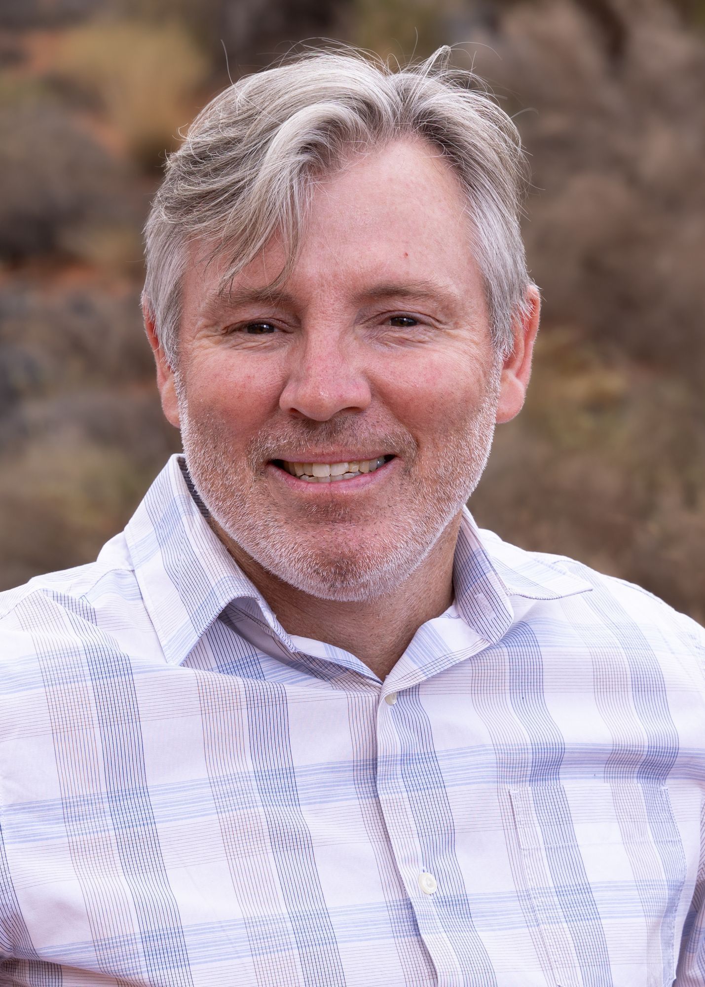 Portrait of a smiling person with gray hair, wearing a white and blue patterned button-down shirt outdoors.