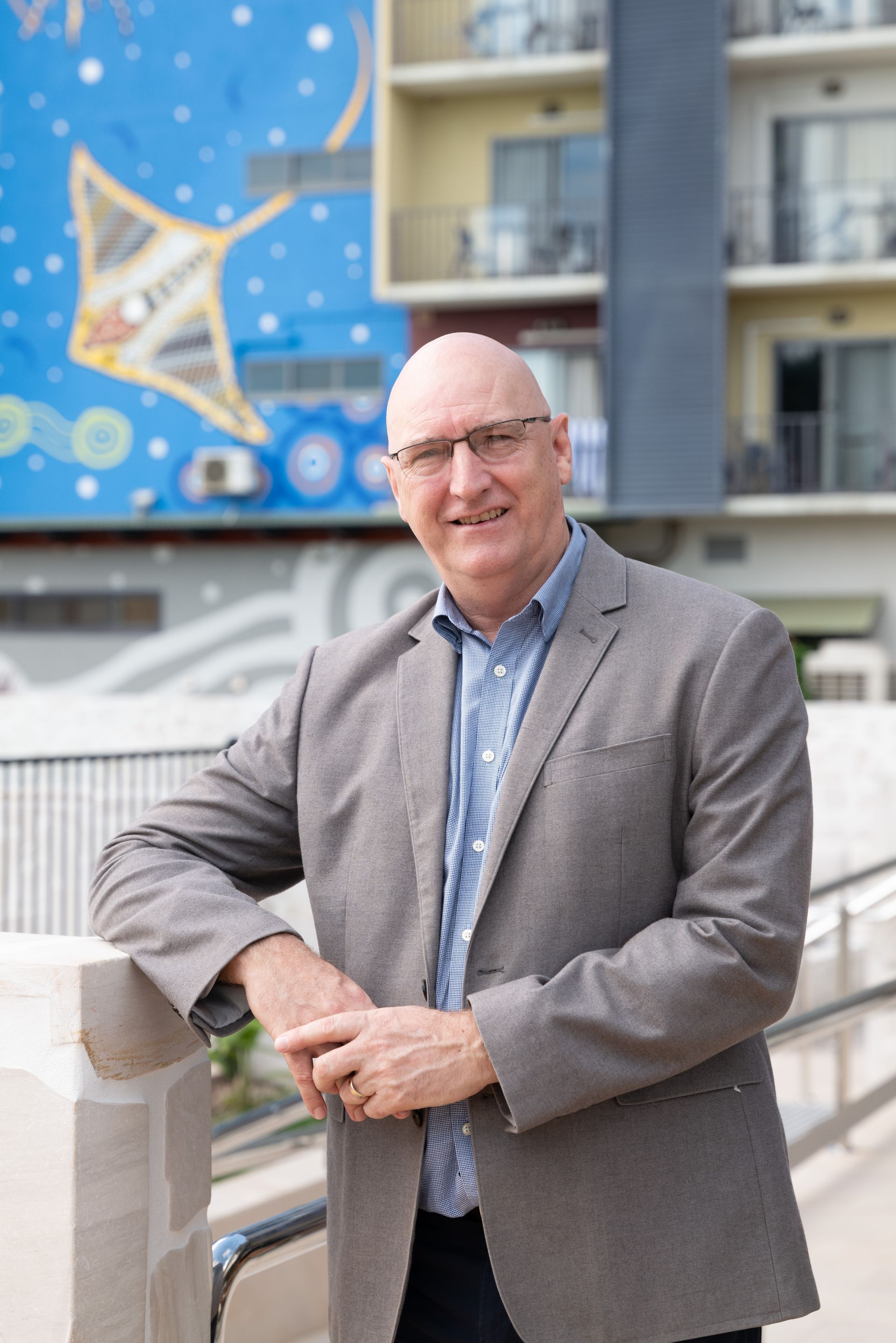 A person in a blazer and patterned shirt smiling, standing on a balcony with a colorful stingray mural in the background.