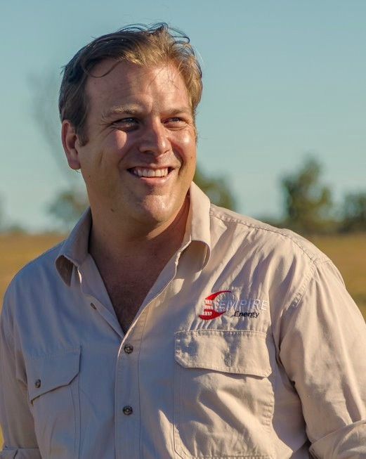 A smiling person wears a tan collared shirt with a red logo, standing outdoors in a sunlit, grassy field.