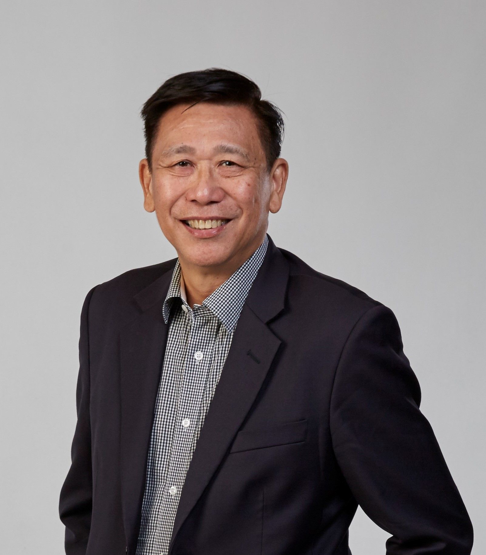 A smiling person wearing a dark blazer over a patterned button-down shirt against a plain, light gray background.