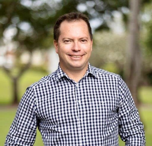 A smiling person wearing a blue and white checkered button-down shirt, standing outdoors with a soft-focus garden background.