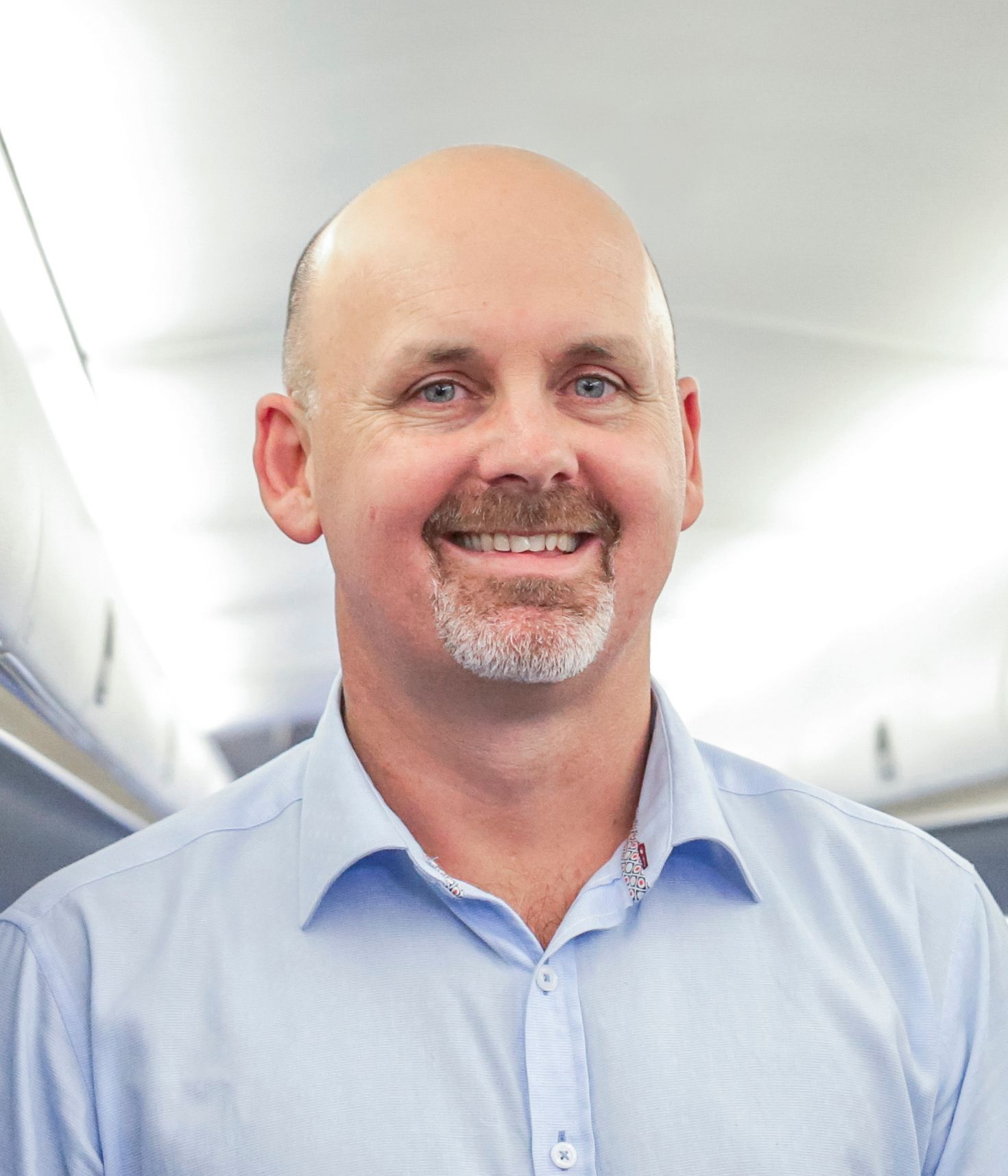 A smiling person with a shaved head and a goatee, wearing a light blue button-down shirt, set against an indoor backdrop.