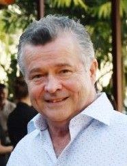 Smiling person in a light blue patterned shirt outdoors with soft, blurred natural background.