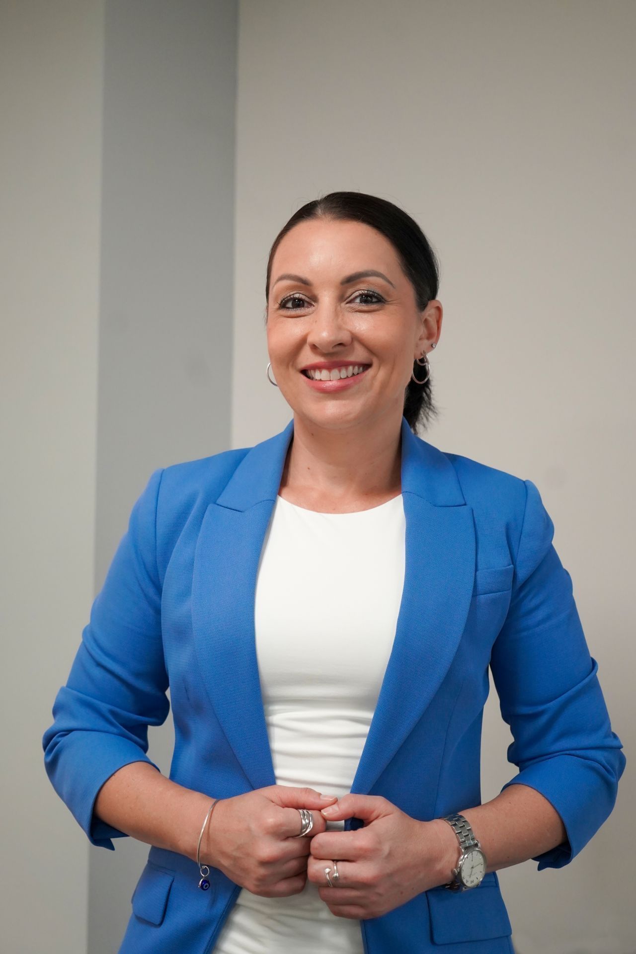 A smiling person wearing a blue blazer over a white shirt stands in front of a plain wall, hands clasped together.