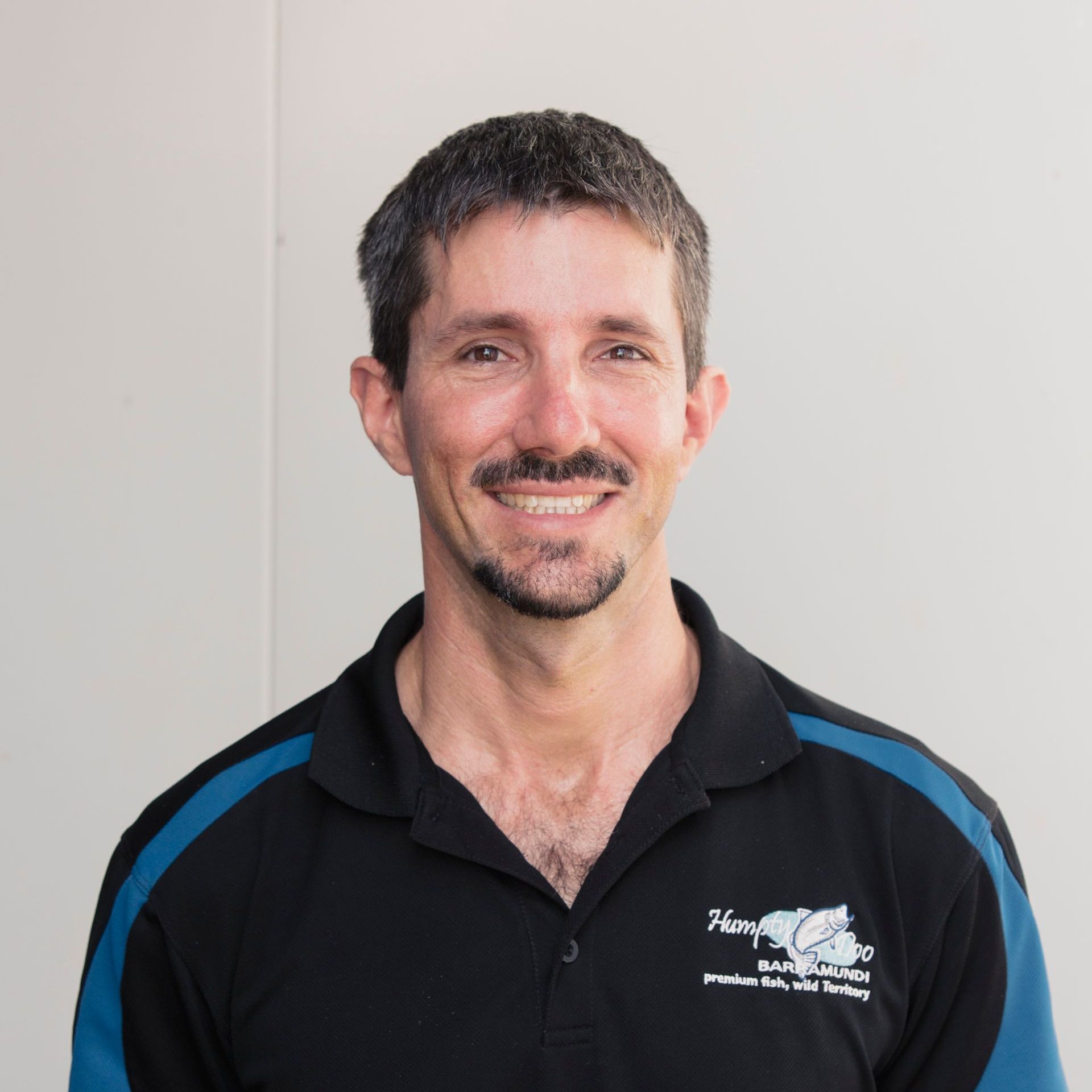 A smiling person wears a black polo shirt with blue sleeve accents and a small logo on the chest, against a white wall.