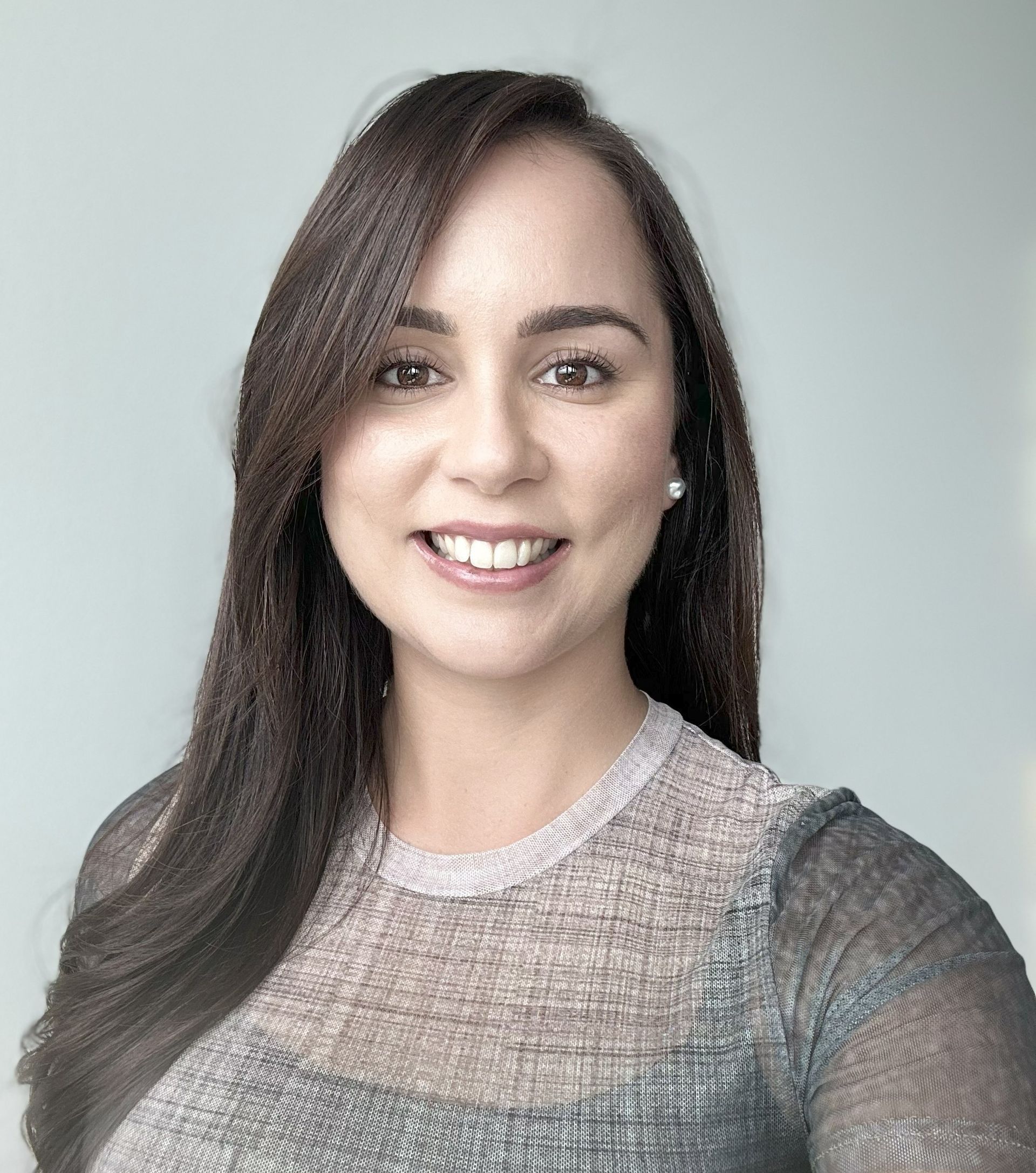 A smiling person with long dark hair, wearing a textured grey shirt against a plain, light blue-grey background.