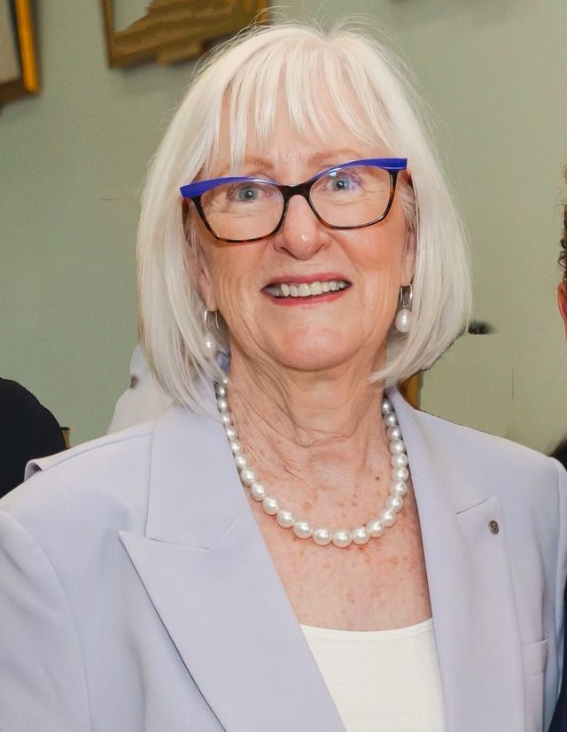 A person with light hair wearing a pale blue blazer, a pearl necklace, and blue-framed glasses smiling at the camera.