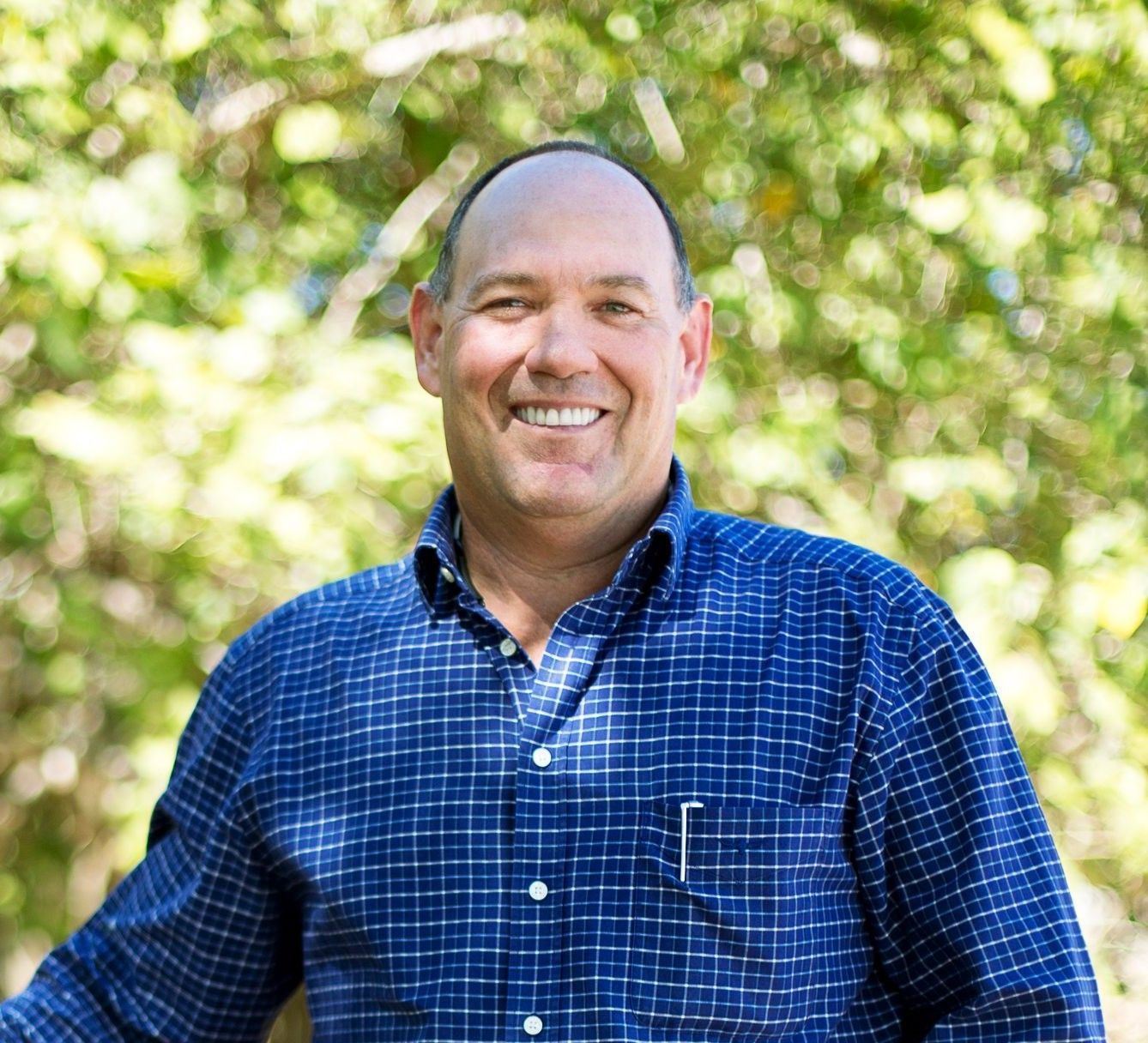 A smiling person with a shaved head wears a blue plaid button-down shirt outdoors in front of out-of-focus green foliage.
