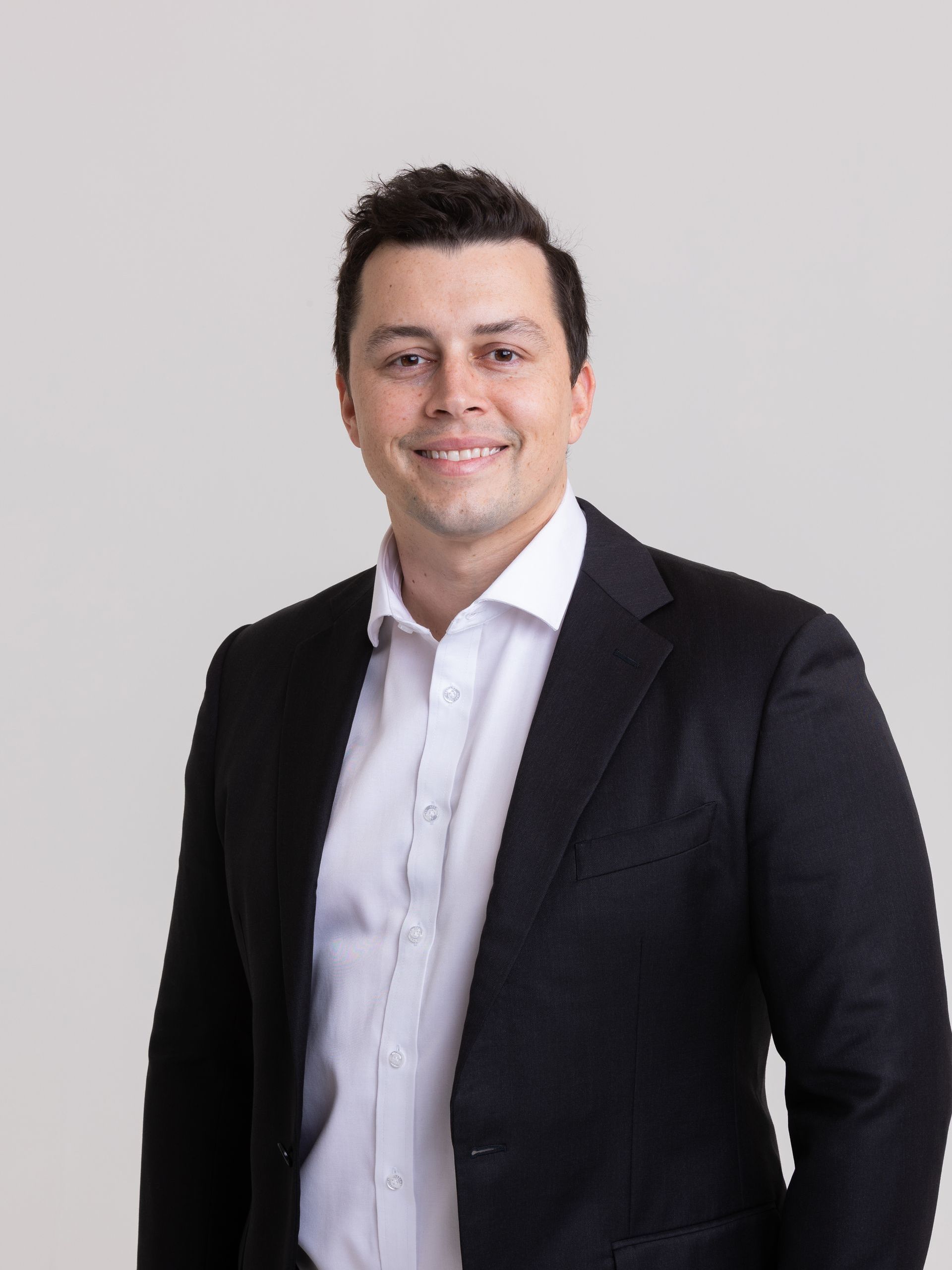 A person smiling in a professional headshot, wearing a white collared shirt and a dark blazer against a grey background.
