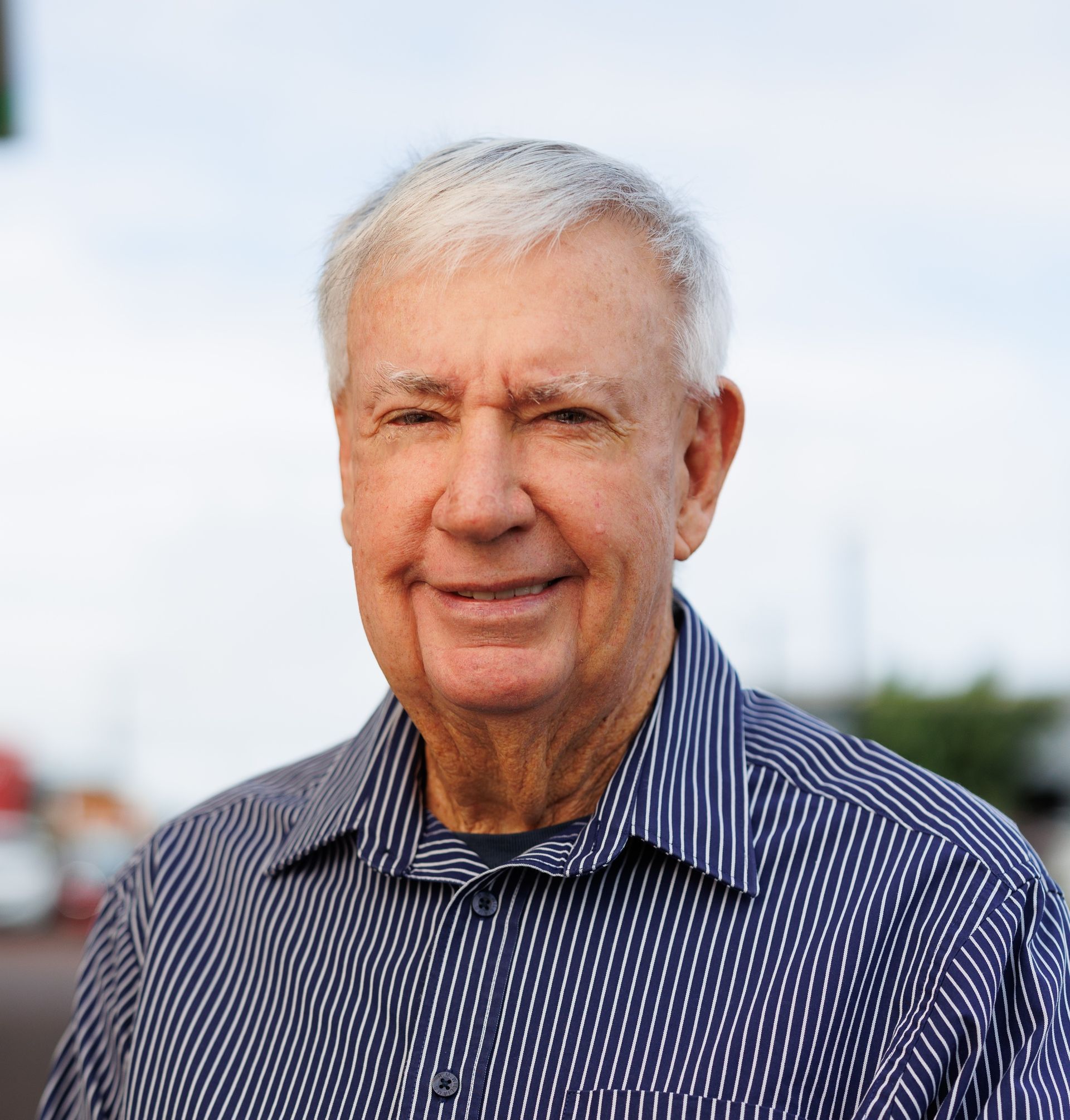 A smiling individual with light hair wears a blue and white pinstriped button-down shirt outdoors.