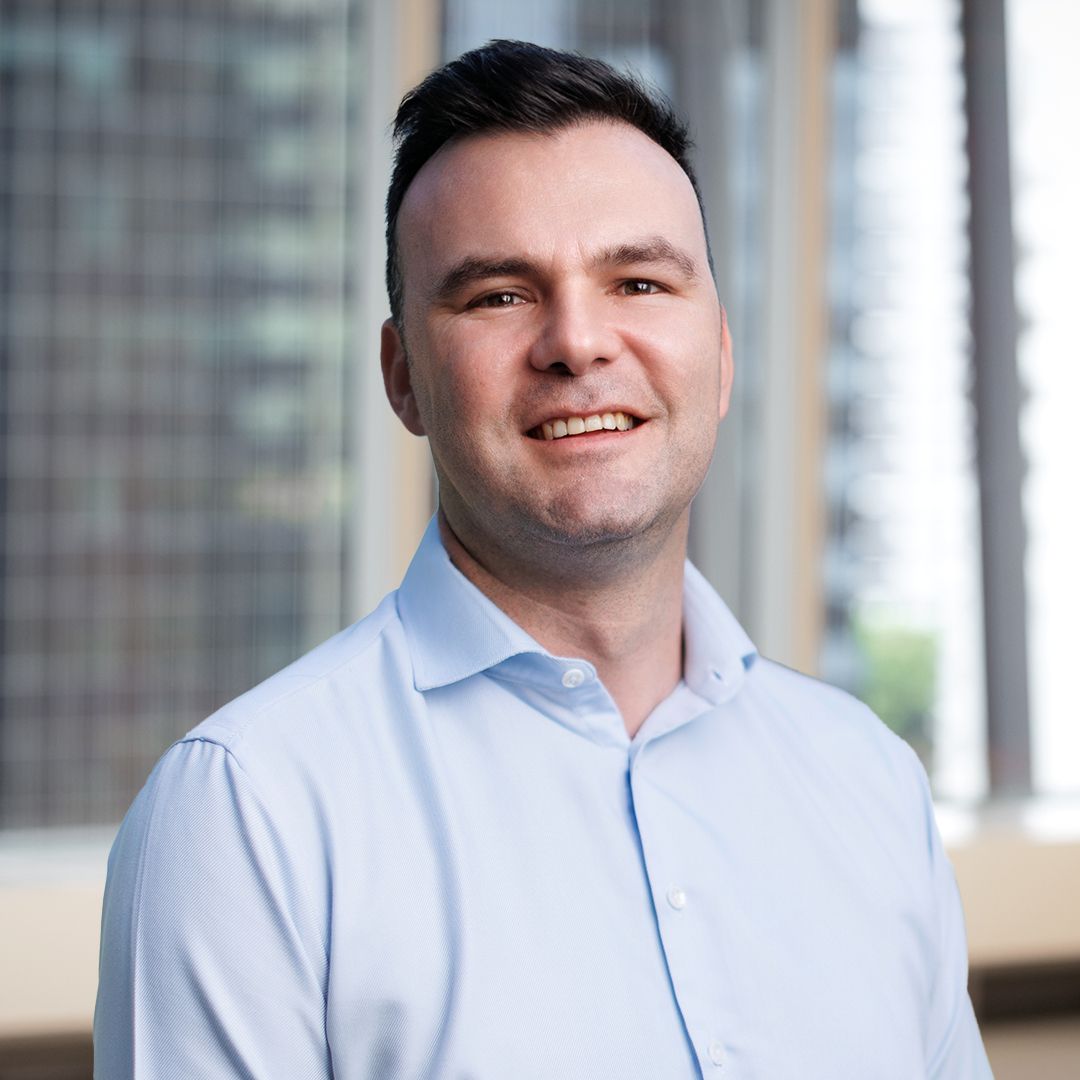 A person wearing a light blue collared shirt smiling warmly in a bright, modern office with windows in the background.
