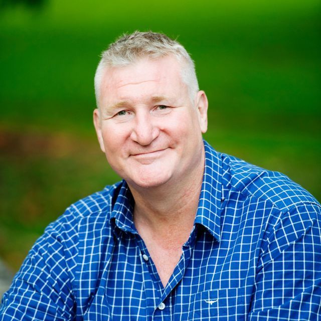 A person with short, light hair wearing a blue and white checkered shirt, smiling softly against a blurred green background.