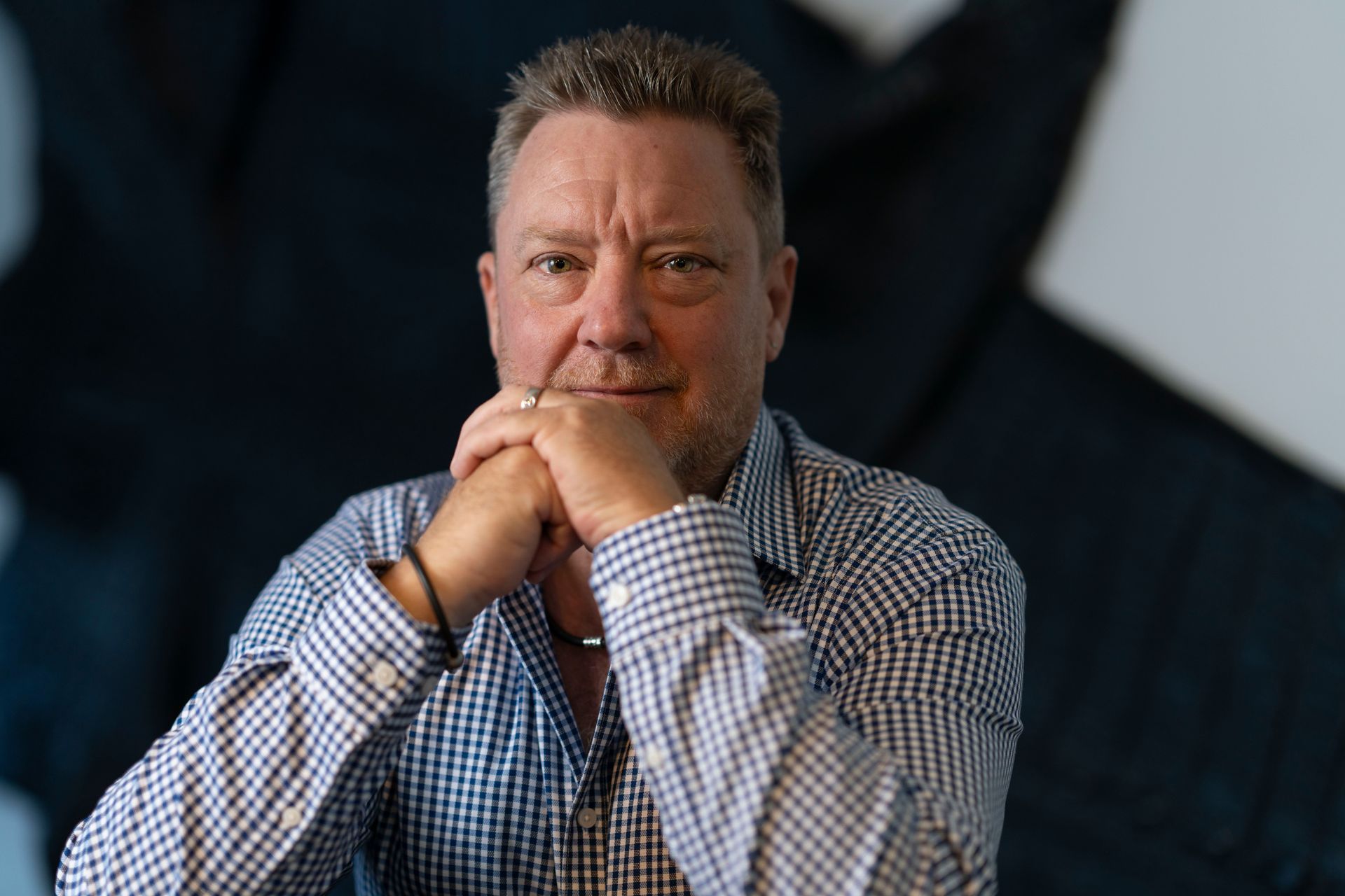 A man in a blue and white checkered shirt with his hands folded under his chin, looking directly at the camera.
