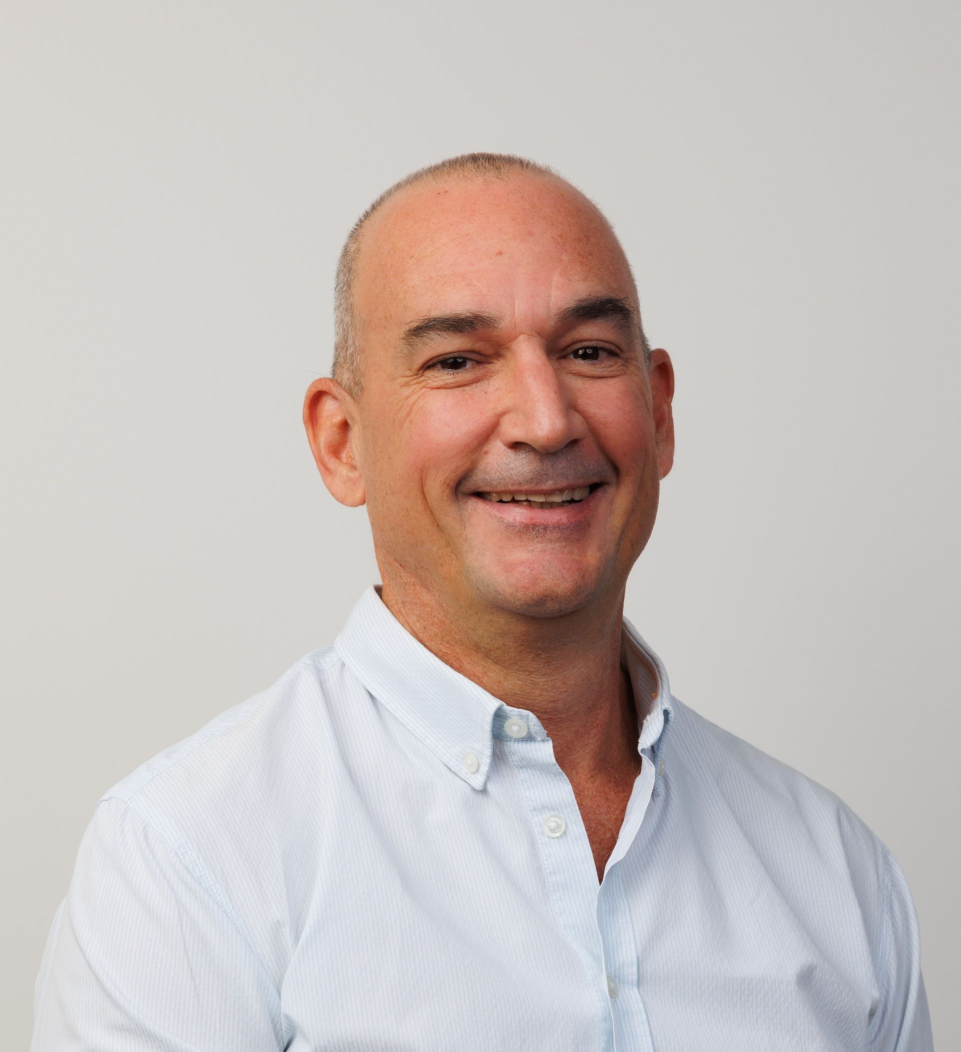 A person with a shaved head wearing a light-colored, button-down shirt smiling against a plain white background.
