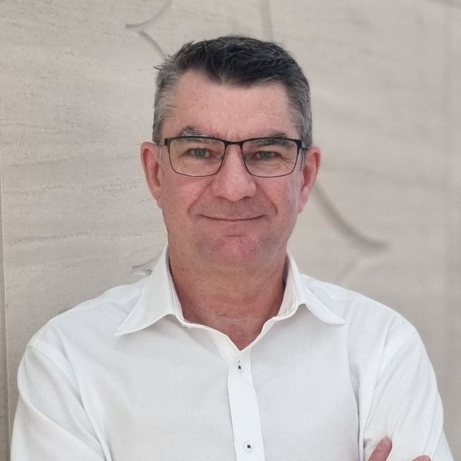 A person with short dark hair and glasses wears a white collared shirt, standing against a neutral textured wall.