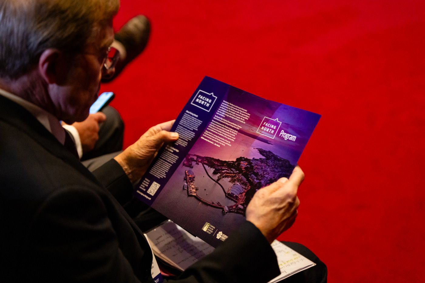 A person in a suit sits in a room with a bright red floor, reading a blue brochure featuring a dark landscape image.
