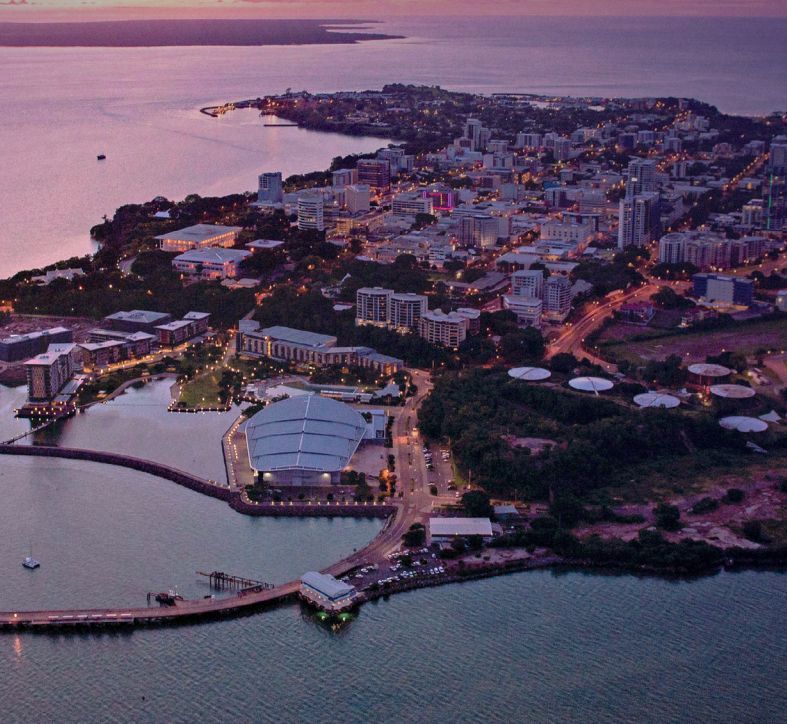 Aerial view of a coastal city at twilight, featuring a large waterfront convention center and illuminated harbor lights.