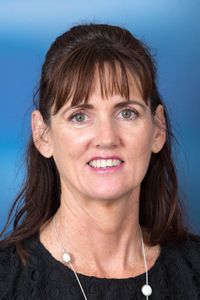 Professional headshot of a person with long brown hair, smiling, wearing a black shirt against a blue background.