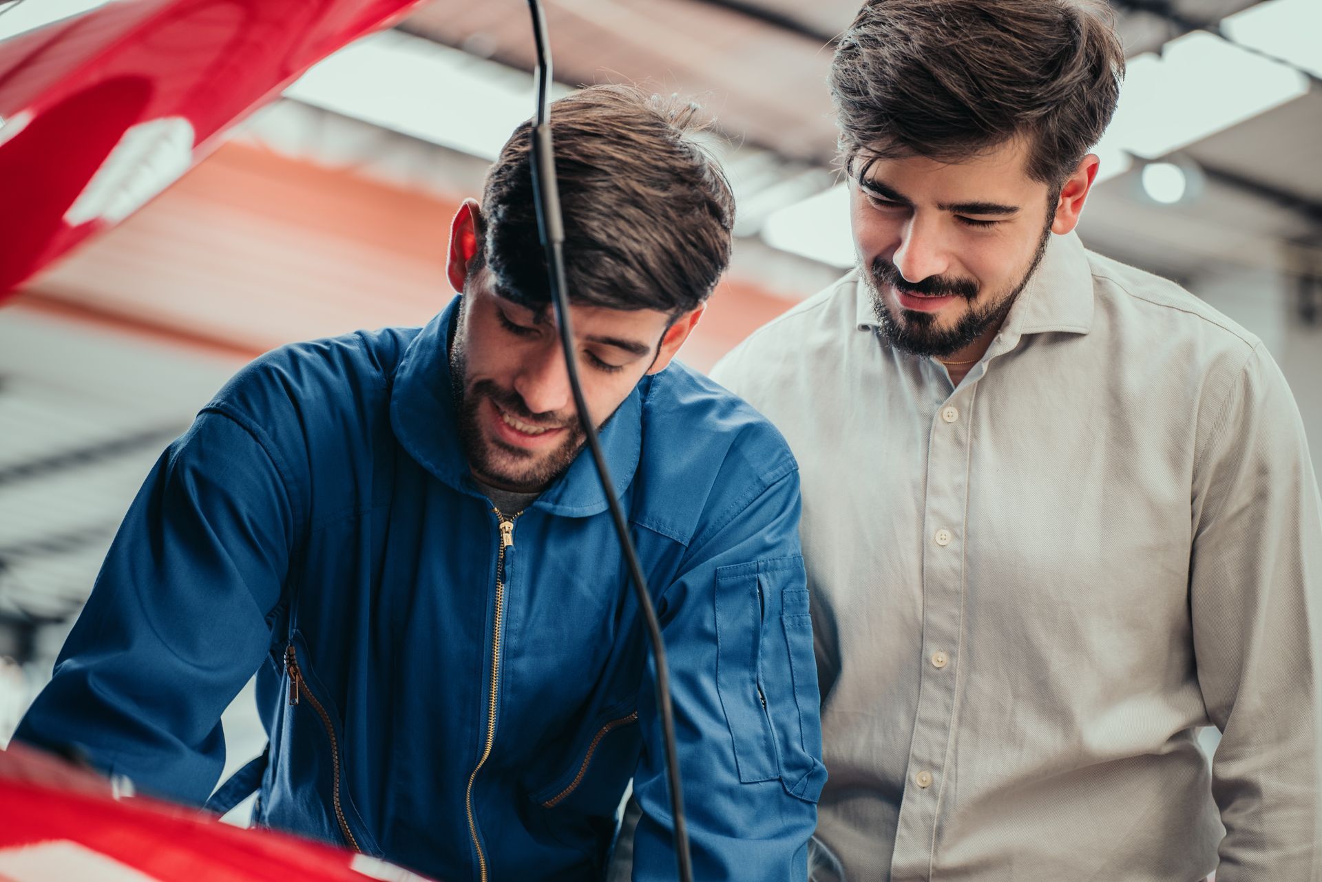 Mechanic in blue coveralls and customer looking at a car engine. Workshop setting, both smiling.