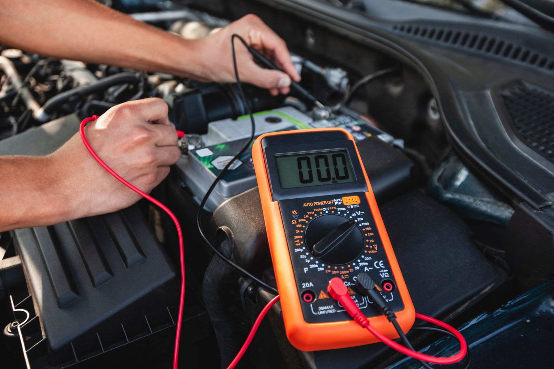 Hands using a multimeter to test a car battery in an open engine compartment.