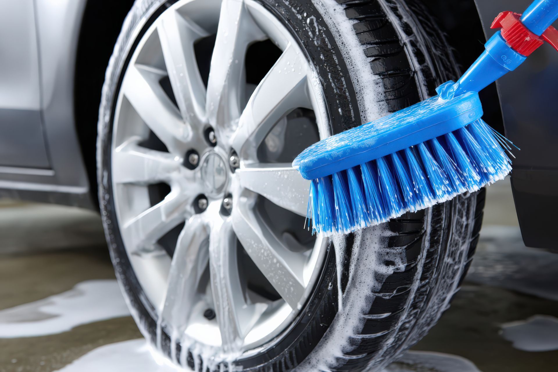 A person scrubbing a car tire with a blue brush covered in soapy suds.