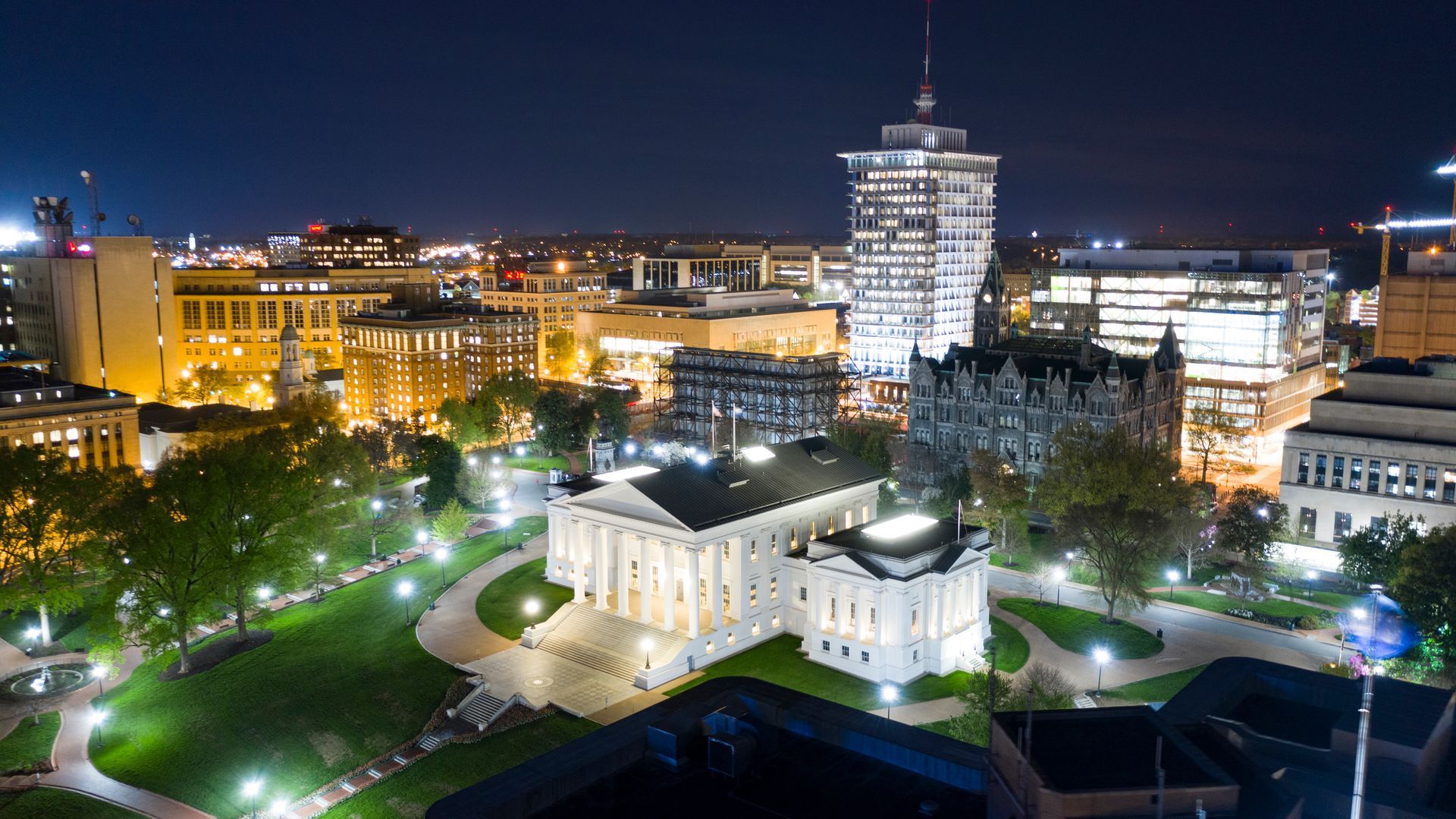 Night view of a city with a lit white building and skyscrapers. Lights illuminate buildings and green spaces.