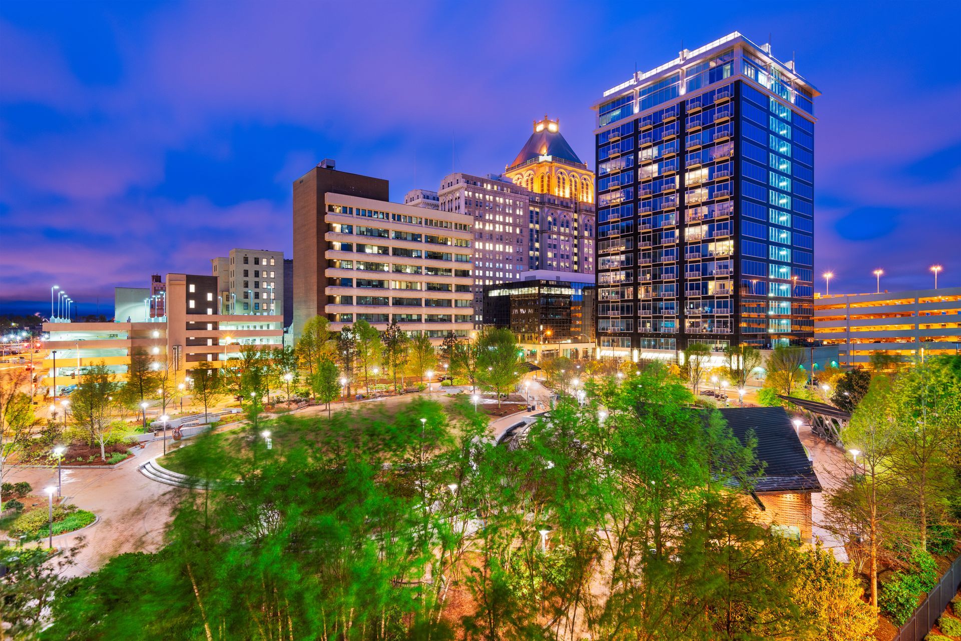 Downtown cityscape at dusk, lit buildings against a deep blue sky; green trees and park in foreground.