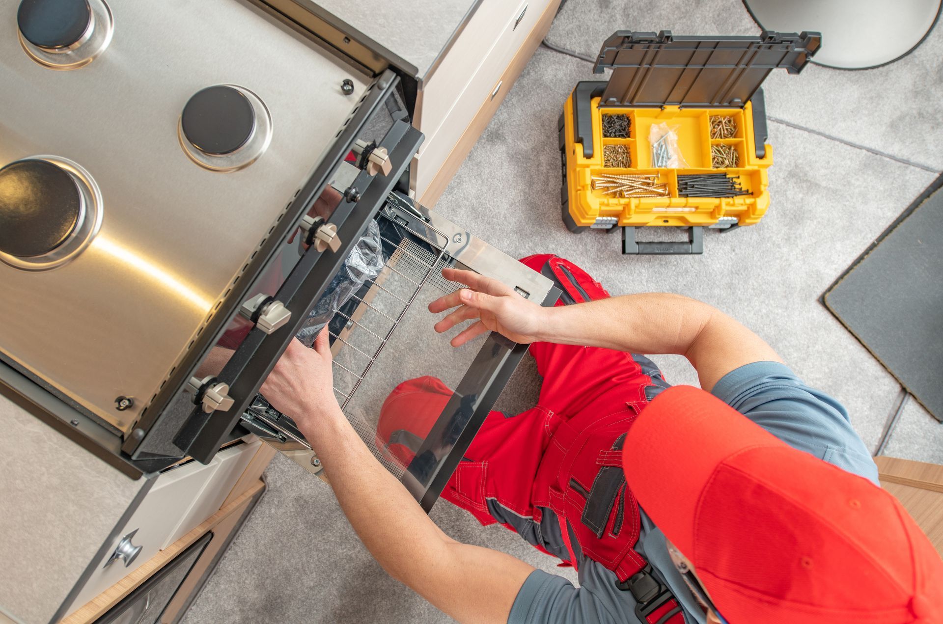 Person in red overalls and hat repairs a gas range in a kitchen, with a toolbox nearby.