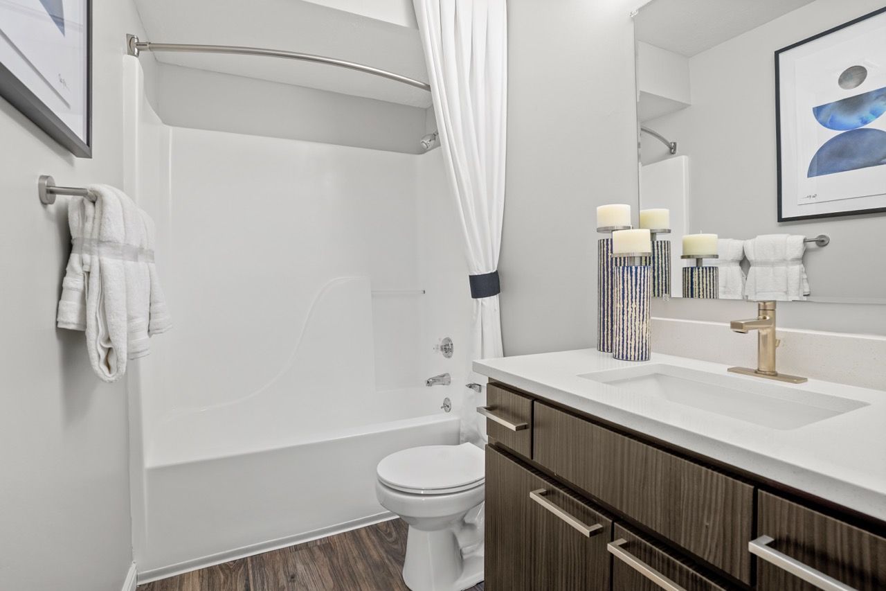 Modern apartment bathroom with a tub/shower, white curtain, towel rack, and wood vanity. Schedule your visit to Turnberry Apartments and discover inviting pet-friendly townhomes in Millcreek, UT, designed for comfortable living.